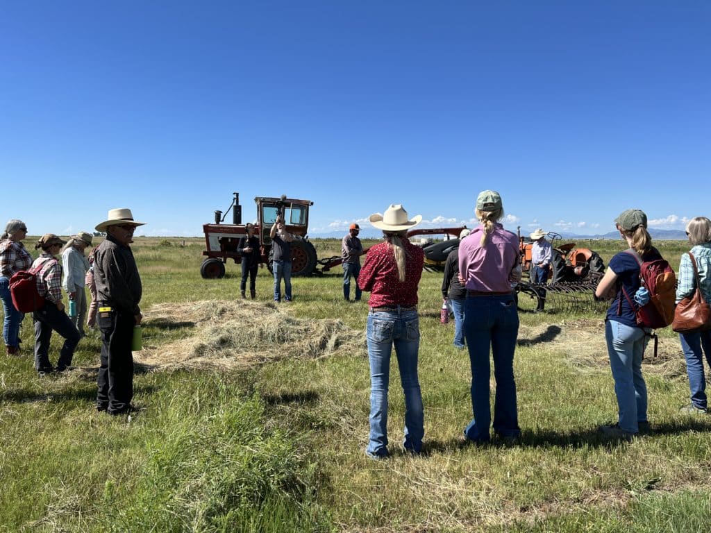 A crowd of farmers and tour attendees watches a presentation in a field next to a tractor and piles of hay.