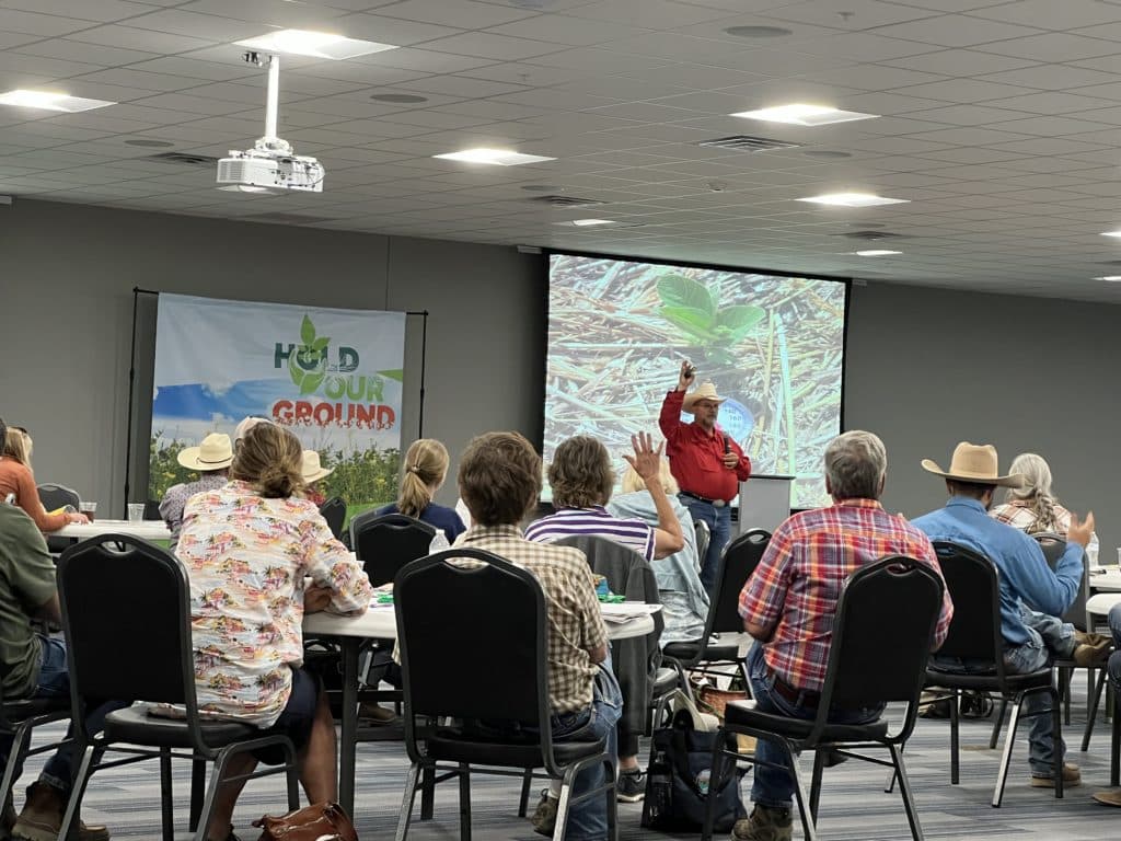 Oklahoma farmer Jimmy Emmons raises his arm in front of the audience, with several members in the audience also raising their hands.